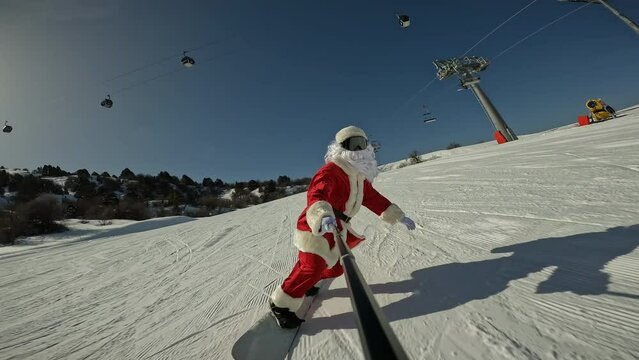 Male snowboarder dressed as Santa Claus snowboarding having fun on ski resort slope with selfie stick. Sliding down mountain trails during Christmas or New Year holidays. Winter extreme sports.