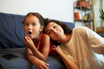 Portrait of cute surprised little girl lying on blue sofa next to smartphone with blank copy space while her pretty mom resting her head on her shoulder to relax and express love after working day