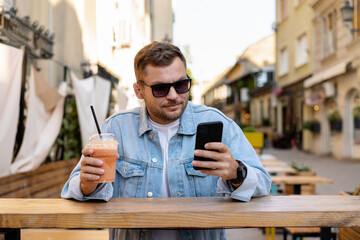 Urban young adult man relaxing in sidewalk cafe on city street, drinking juice and using his smart...