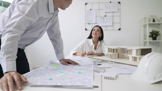 Skilled caucasian interior designer working with house model while discussing with asian architect engineer about house plan on meeting table with blueprint and house model scatter around. Immaculate.