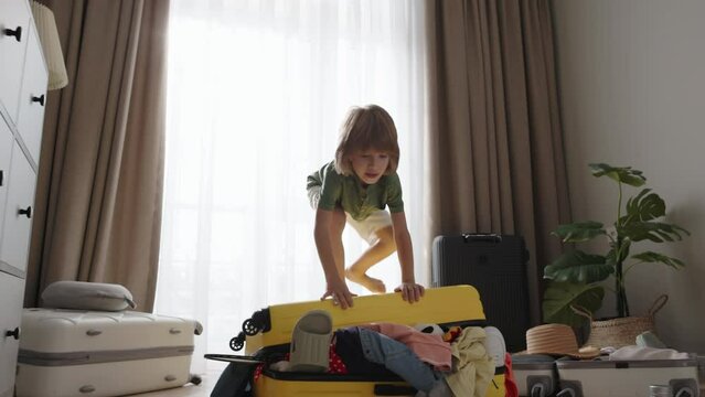 Throwing another toy into the suitcase and trying unsuccessfully to close it, boy dances joyfully on the floor ready for the trip. Child's excitement fills the air as he jumps on top of yellow luggage