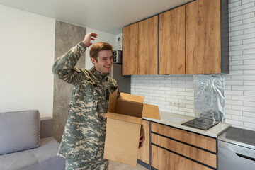 Smiling man in camouflage holding cardboard box in new apartments