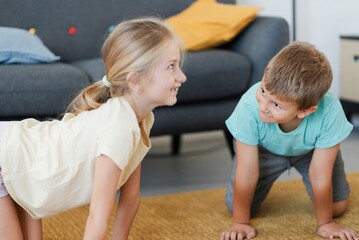 Cheerful little girl and boy in casual clothes practicing Chakravakasana yoga pose on carpet in living room at home