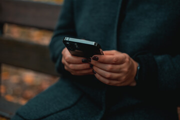 Autumn Smartphone Use: Close-up of hands delicately holding a smartphone, outdoors on a park bench in autumn.