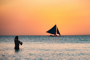 (Selective focus) Stunning view of a boat sailing during a beautiful sunset in the background and the silhouette of a blurred person swimming in the foreground. White Beach, Boracay, Philippines.