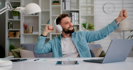 Handsome young bearded man wearing casual clothes listening music in wireless headphones sits the...