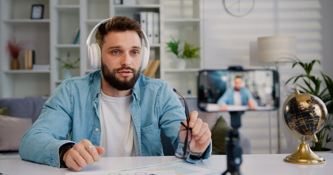 Focus on mobile phone camera from which good-looking confident in headphones adult man recording video vlog for his internet channel from modern light home room, indoors