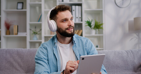 Attractive man wearing headphones sit on sofa holding tablet device while having a video call on mobile tablet screen talking. E-commerce, virtual services concept