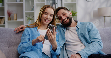 Smiling young woman showing photo video content in social network to husband, using smartphone. Happy family couple using modern technology gadget together sitting on sofa at home - obrazy, fototapety, plakaty