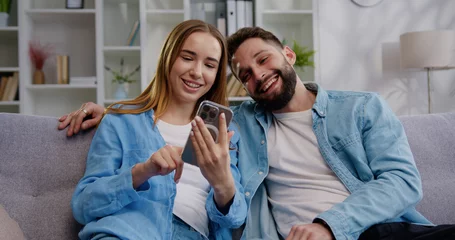Foto op Canvas Smiling young woman showing photo video content in social network to husband, using smartphone. Happy family couple using modern technology gadget together sitting on sofa at home © serg