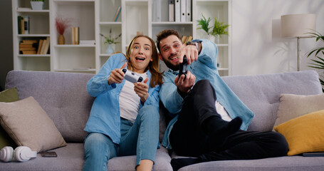 Happy couple sitting on the sofa playing video games, using controllers. competitive Girlfriend and boyfriend in love have fun playing in online video games in living room at home together - obrazy, fototapety, plakaty