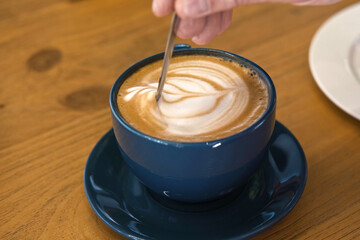 Hand is Mixing the Milk Foam of Latte. Person Stirring Cappuccino with Steel Spoon. Hot Coffee in a White Classic Ceramic Cup on Wooden Table. Modern Coffee shop, Cafeteria. Thick Froth. Close-up View