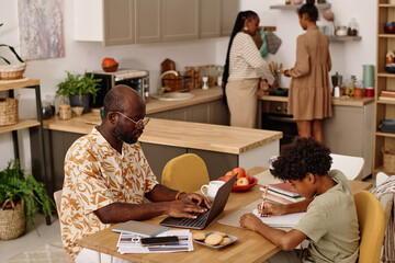 Father working on laptop, son doing homework when mother and daughter cooking dinner