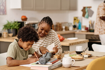 Obraz na płótnie Canvas Smiling Black woman helping preteen son with doing homework for upcoming class