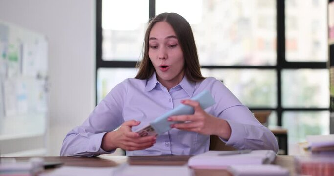Young woman opens envelope containing big banknotes. Female reacts positively to money by dancing at table. Joy of receiving cash slow motion
