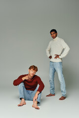 red haired man in sweater squatting on floor with african american man standing on gray background