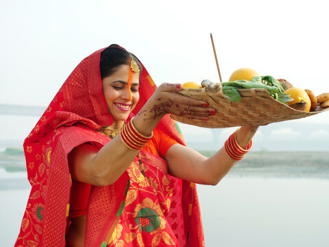Shot of a woman posing for the camera while standing in a river - Chhath Pooja celebration  praying to Sun. A woman dressed in a traditional Chhath pooja attire while holding the prasad in a bamboo...