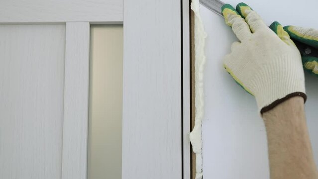 Cut off excess hardened polyurethane foam with a knife after foaming the cracks in the door frame. Construction and repair. Installation and fastening of a wooden frame.