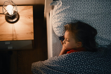 Child sleeping in bed at late night. Cute toddler girl pillow covered with warm blanket. Kid having...