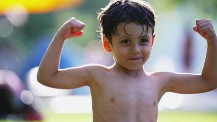 One adorable little boy flexing muscles sitting on grass outside after swimming at the pool during warm summer day. Child shows tiny biceps © Marco