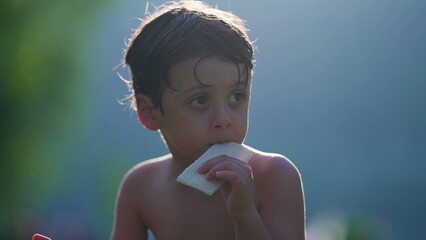 Pensive child eating biscuit while drying in the sun after playing at pool. Shirtless small boy enjoys summer vacations observing surroundings