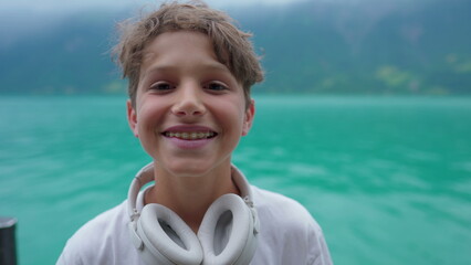 Portrait of handsome young boy removing headphones and smiling to camera while standing in lake...
