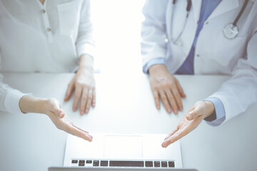 Doctor and patient sitting near each other at the table in clinic. The focus is on female hands pointing into tablet computer touchpad together. Medicine concept - obrazy, fototapety, plakaty
