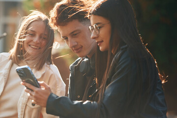 Girl is showing something in smartphone. Three young students are outside the university outdoors