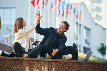 Giving high five, success. Two young students are sitting outdoors against university