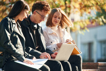 Learning, sitting and using laptop. Three young students are outside the university outdoors