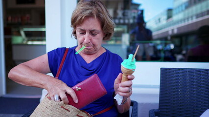 Senior woman putting leather wallet away inside purse while holding waffle ice-cream cone in parlor...