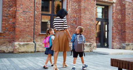 Children with backpacks hold their mother hand. Happy family mom and children go to school. Mother leads her son and daughter by hand through school yard. children go to class holding mother hand.