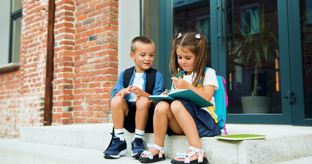 Portrait of attractive couple of little friends boy and girl sitting on stairs by school building in smart casual style, writing notes outside together smiling. Outside. Educational school program for