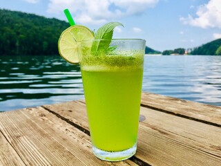 Glass of mint lemonade with a slice of lemon placed on a wooden pontoon near the lake in summer