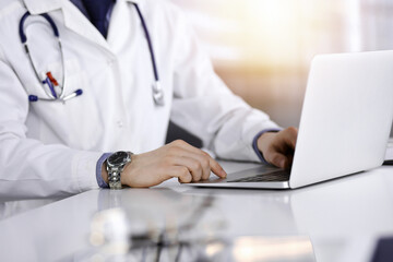 Unknown male doctor sitting and working with laptop in a darkened clinic, glare of light on the background, close-upof hands