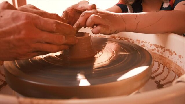 Potter teaching little girl to craft ceramic mug on a pottery wheel outdoors at warm sunny day	