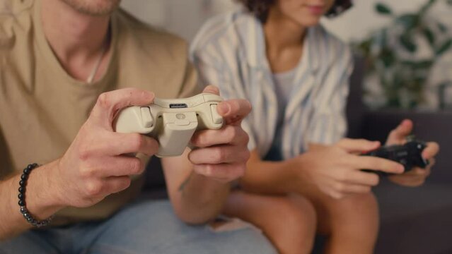 Selective focus medium close-up shot of game controllers in hands of young woman and man sitting on couch