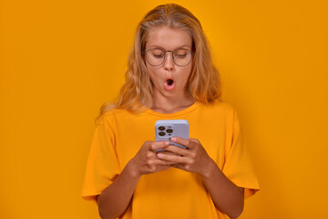 Young shocked cute Caucasian woman teenager using smartphone opens mouth and is surprised after reading news in tabloids about personal lives of stars stands posing in yellow studio. Mobile phone, app