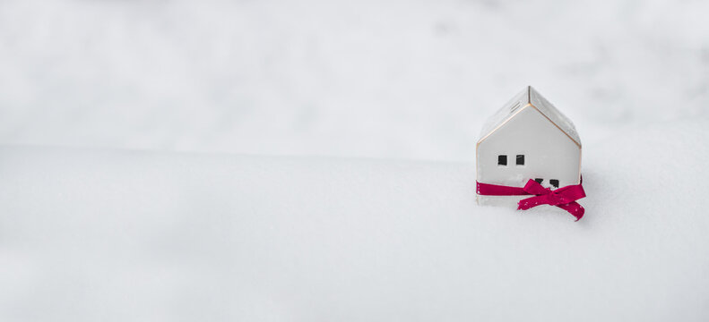 Merry Christmas and 2024 happy new year horizontal banner with small toy model house wrapped in red satin ribbon on winter snow background. Miniature white toy house model. High quality photo