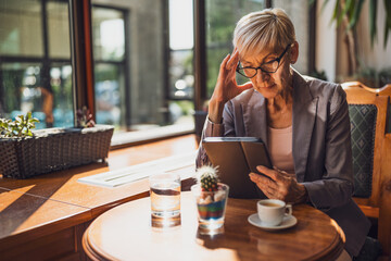 Mature woman is sitting in cafe and relaxing. She is drinking coffee and using digital tablet.