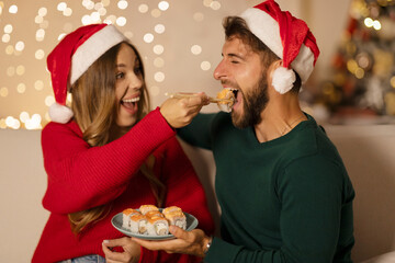 Couple eating sushi together, woman feeding husband, wearing Santa hats and sitting in living room...