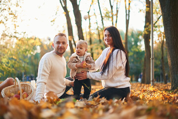 Two parents are sitting with cute little boy in the autumn park on the fallen leaves and butternut squash