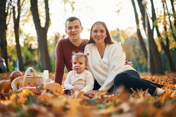 Little daughter with mother and father, that are sitting in the autumn park on the fallen leaves...