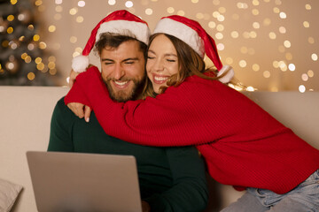 Happy spouses in red Santa hats using laptop woman embracing husband, sitting on sofa in decorated apartment, with glowing garlands and x-mas tree