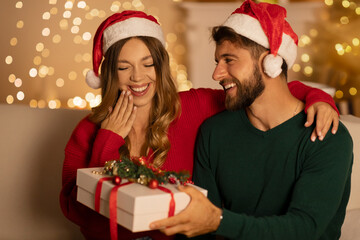 Romance and winter holidays. Surprised woman looks at gift box from husband, sitting in living room interior with Xmas tree with luminous garlands