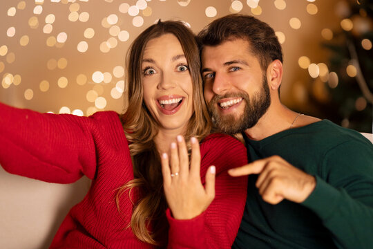 Happy couple got engaged on Christmas evening, cheerful man and woman taking selfie, showing engagement ring and smiling