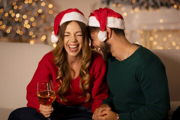 Laughing young european guy and lady in Santa Claus hats hugging in living room with Xmas tree and decor with glowing garlands