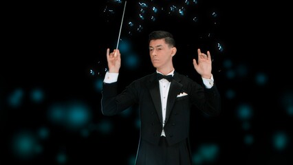 In a close up shot against a black background, a young man in a tuxedo stands in his hands with a wand. Depicts a conductor who controls the notes, they surround him and float. He creates art, music