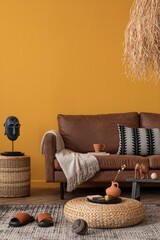 Creative composition of ethnic living room interior with brown sofa, yellow wall, braided pouf, patterned pillow, sculpture, grass lamp, patterned rug and personal accessories. Home decor. Template.