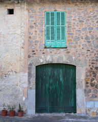 An old wooden green door with an iron handle in a stone wall, at the top there is a window with wooden shutters.